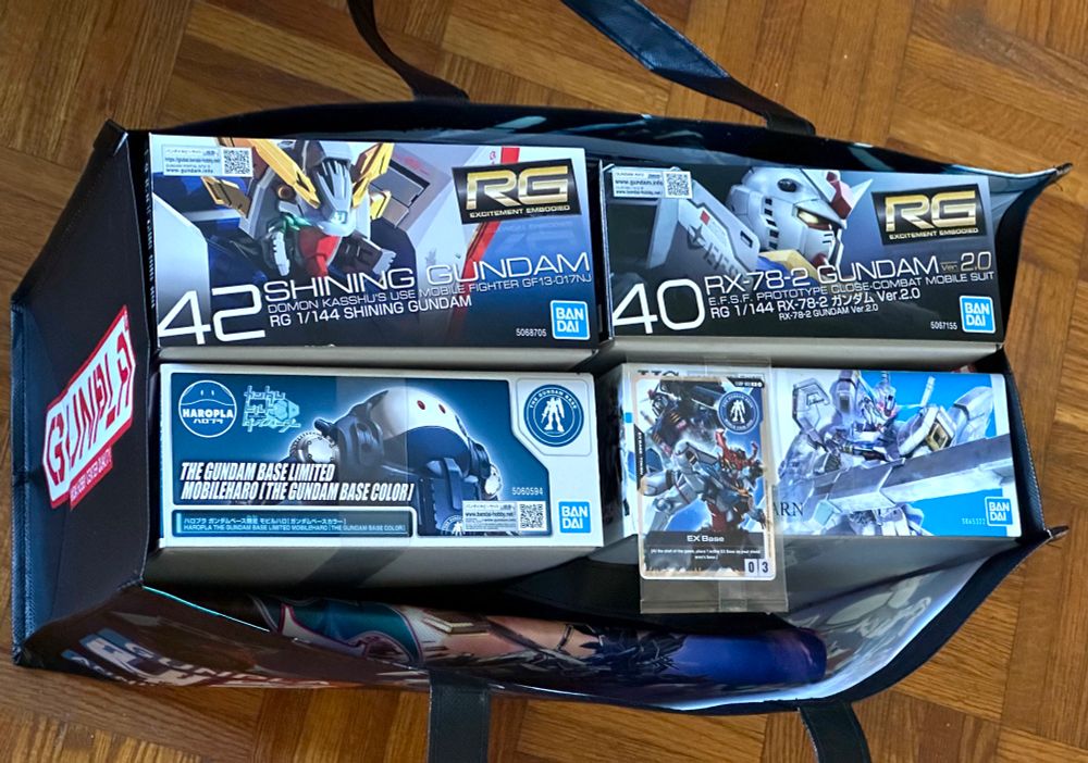 A bag containing four model kits: a Real Grade Shining Gundam, a Real Grade RX-68-2 2.0, a Gundam Base Limited Mobileharo, and a High Grade Gundam Calibarn. A promotional Gundam trading card game is also sitting on top of the boxes.