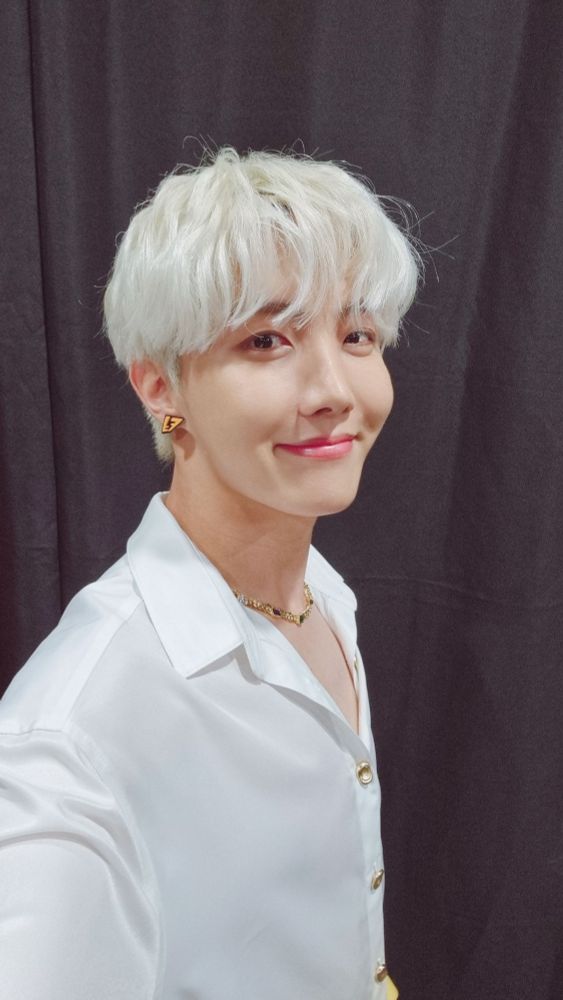 A selfie of j-hope, softly smiling. It seems like he's backstage at a concert. His hair is dyed platinum white and he's wearing a white shirt with gold buttons. It looks like he's glowing, like an elf prince at the end of a fantasy novel.