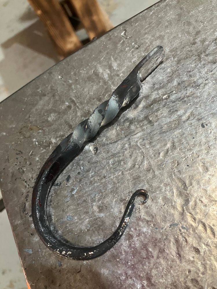 Hand-forged metal hook, just-quenched and still wet. It is lying on a shiny metal base which is the anvil used for forging it. The hook has a tiny scroll end, a generous curve, a twist, and a flattened ‘splat’ at the end that can be drilled for attachments.