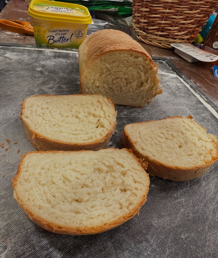 A finished loaf of white bread on a black cutting board. with three slices cut off and a tub of I Can’t Believe It’s Not Butter in the background 