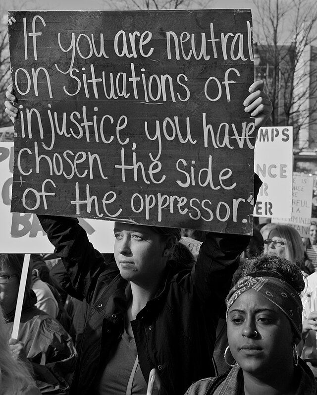 Woman at a protest holding a sign with the Archbishop Desmond Tutu quote, "If you are neutral on situations of injustice, you have chosen the side of the oppressor."