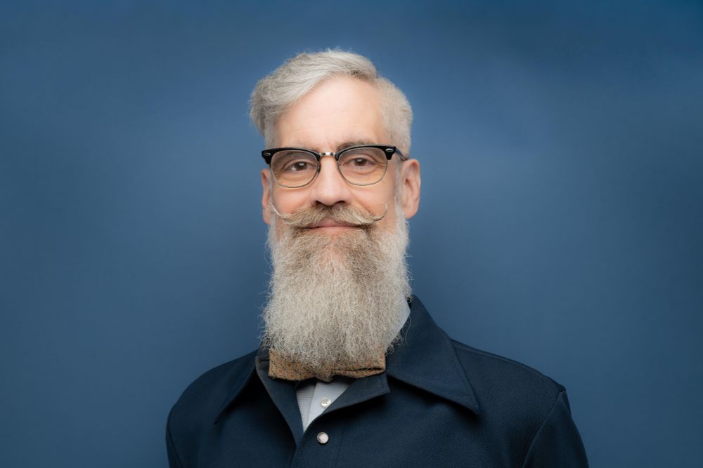 Headshot of Matthew Coomber. Silver hair and beard with curled mustache. Glasses and bowtie.