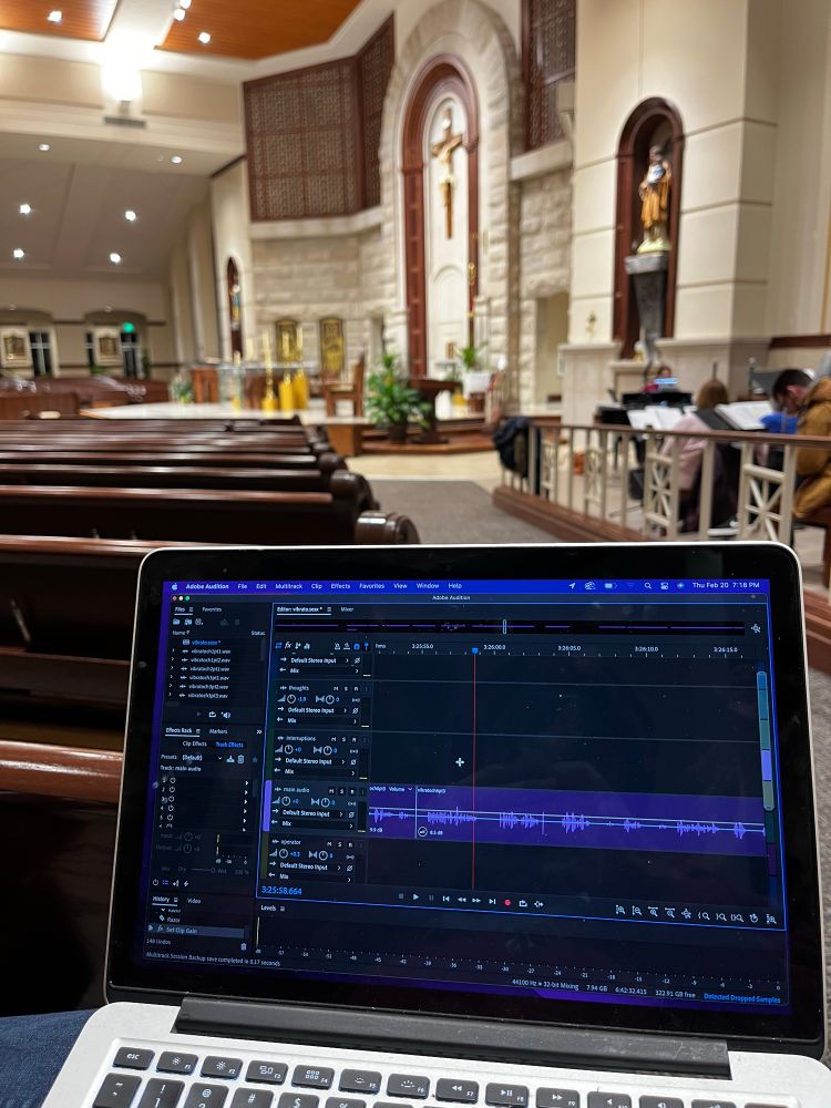 A laptop in the foreground open to Adobe audition, audio editing software, with a multitrack file pulled up. In the background you can see that I am sitting in a church pew looking at the altar with a giant crucifix above it.