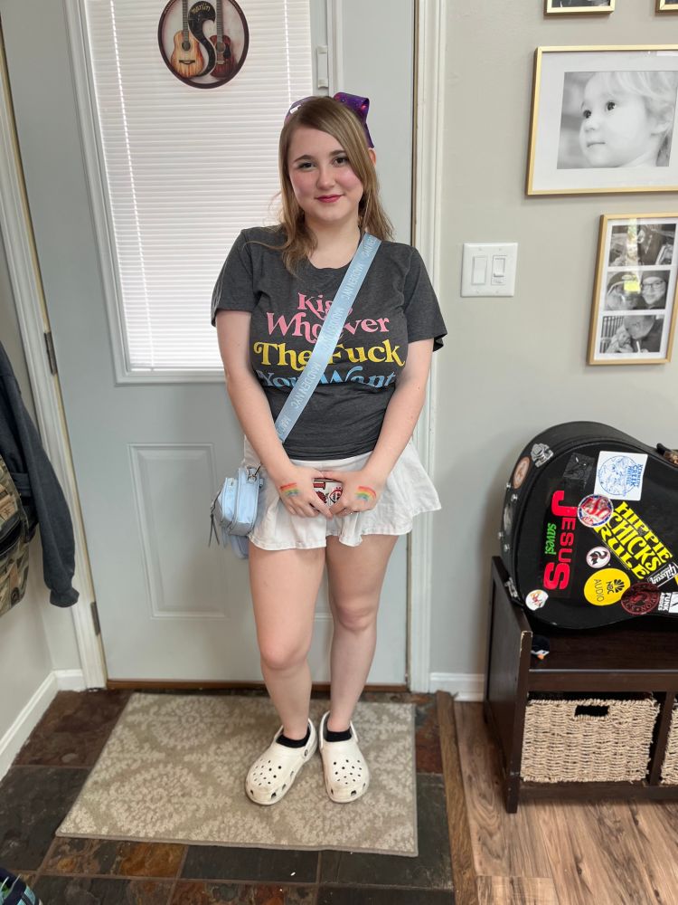 A picture of a teenaged girl wearing a shirt that says “kiss whoever the fuck you want” in pansexual colors