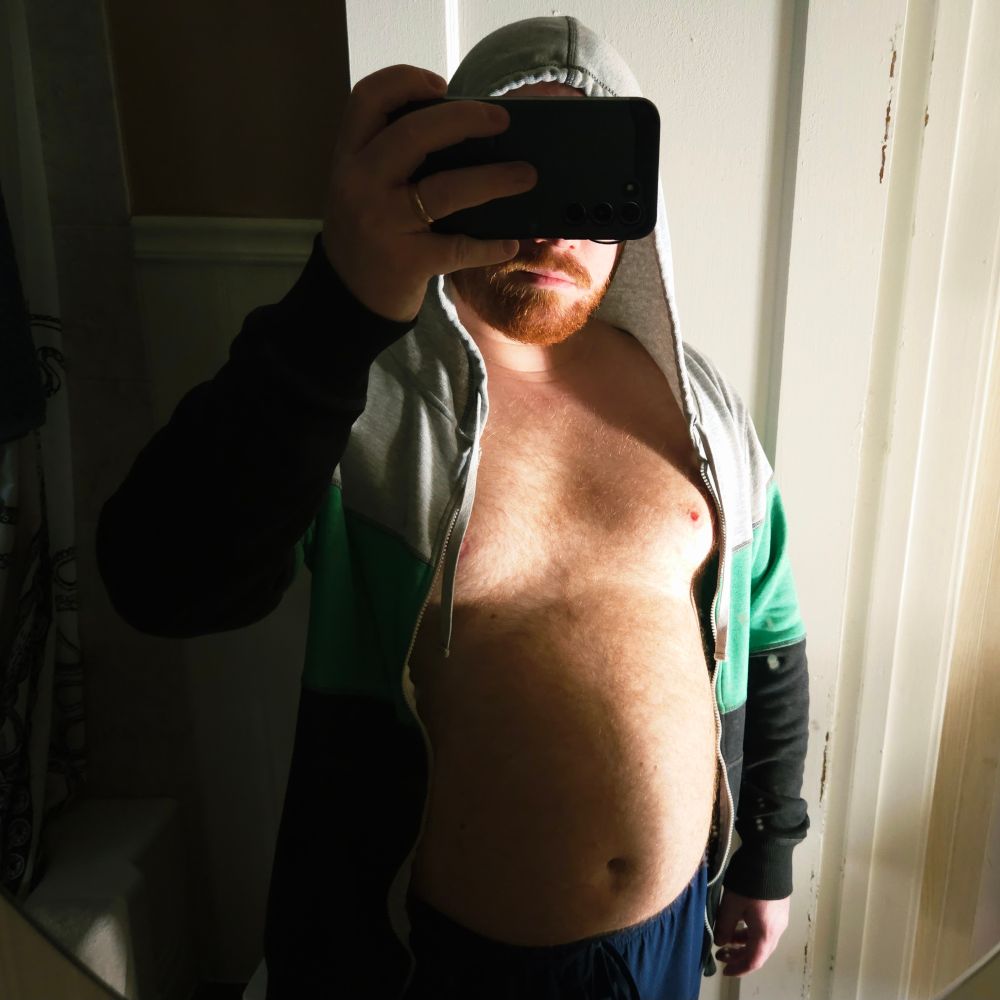 Mirror selfie in a bathroom. A white, bearded, redheaded man with a hairy dadbod and a round tummy is standing with his phone in front of his eyes. He's wearing blue athletic shorts and a grey, green, and black zip-front sweatshirt with a hood on. Light comes through the open door, shading his chest and tummy nicely.
