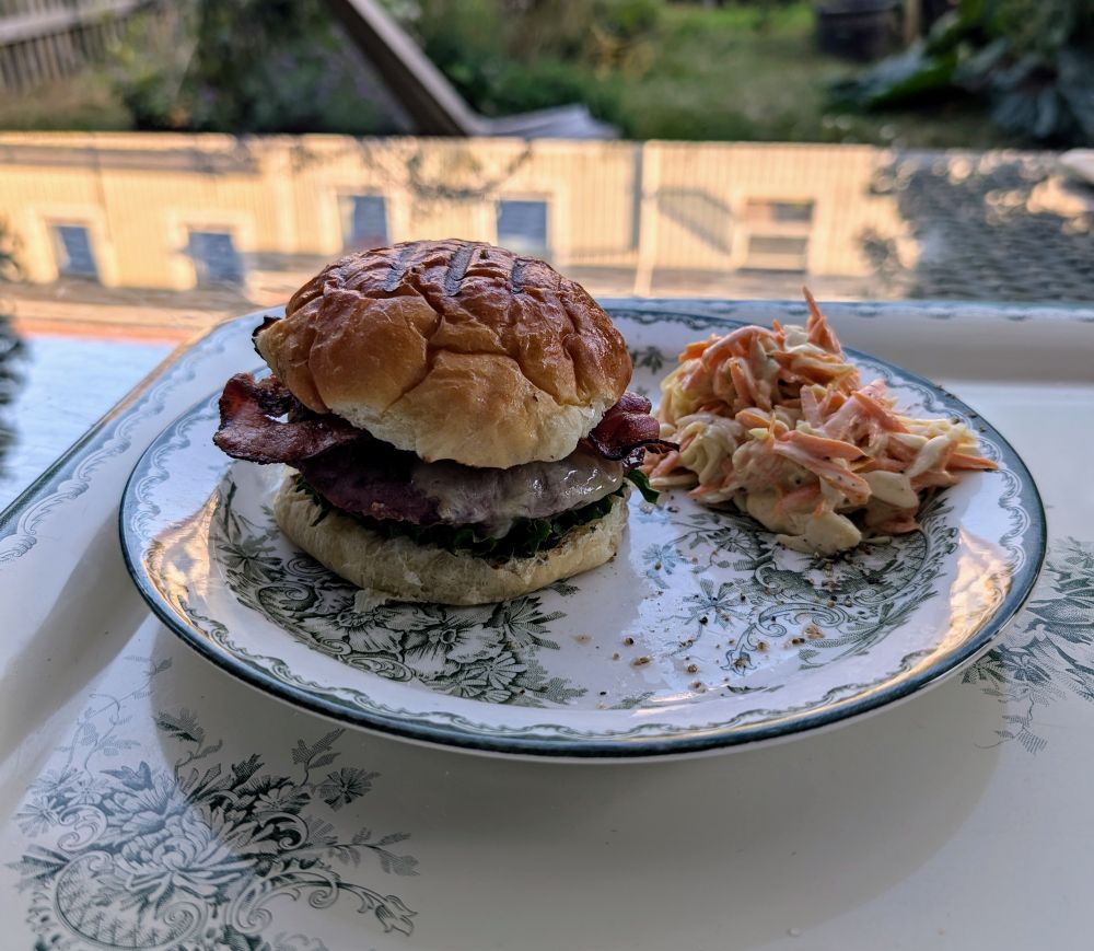 Hamburgare med ost och bacon, coleslaw på sidan. Radhusträdgård i bakgrunden.