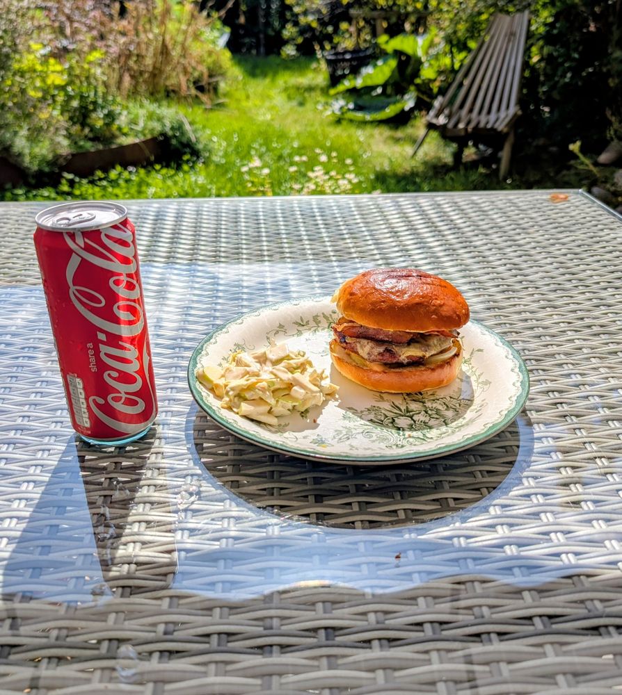 Hamburgare med bacon, ost och lök på en tallrik tillsammans med coleslaw på äpple och vitkol. Brödet har lätta grillränder. Trädgård i bakgrunden och en burk coca-cola bredvid tallriken.