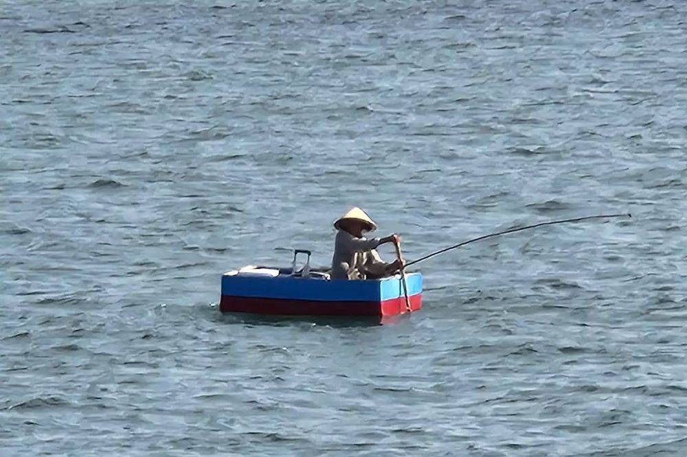 Man with face shrouded and round hat fishes with rod and reel from a red and black box floating on waves