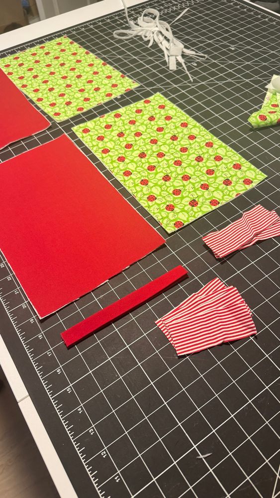 two rectangles of red fabric, two triangles of green fabric with printed leaves and ladybugs, all laying on a black cutting mat. 