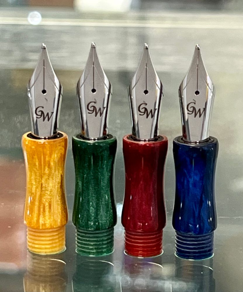 Four fountain pen sections, one yellow, in green, one red, and one blue, are shown with steel nibs sitting upright on a glass surface. 