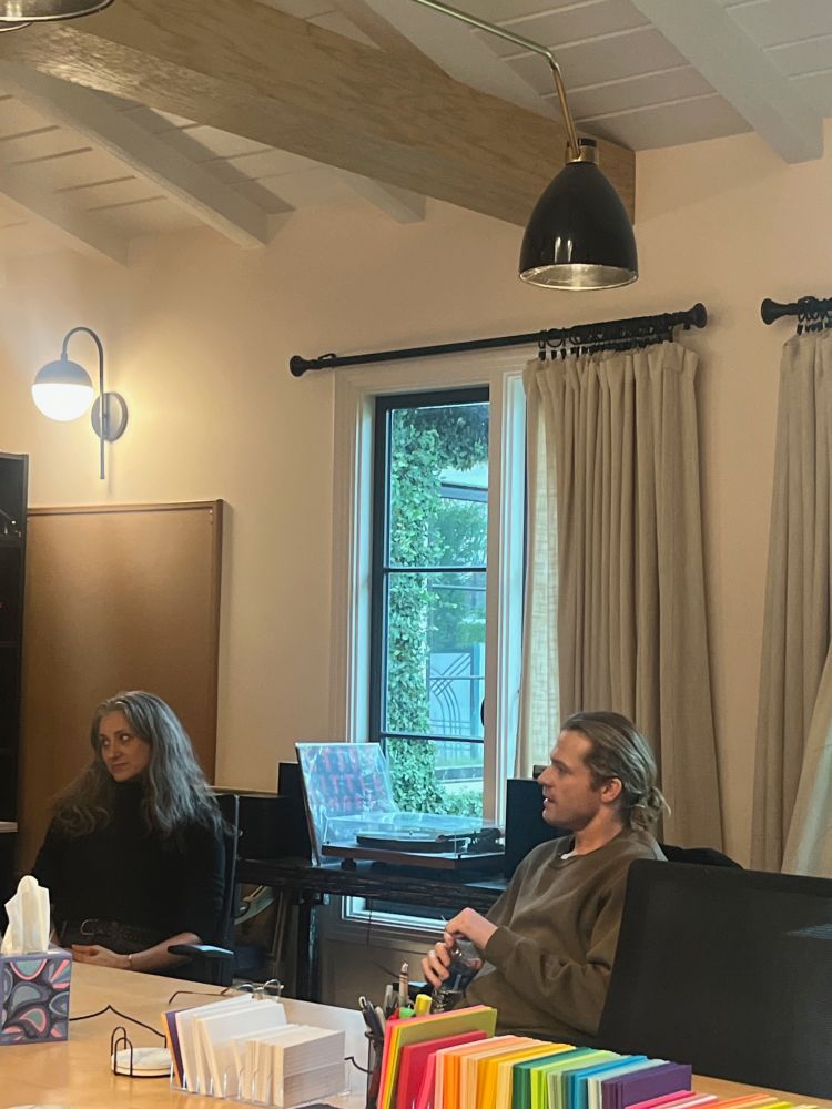 Sam Reid holds a water bottle at the Writer's Room writing table. Exec Producer Hannah Moscovitch sits next to him. They both look at Rolin Jones, showrunner, off-camera. There is a line of white and colored notecards on the table. 