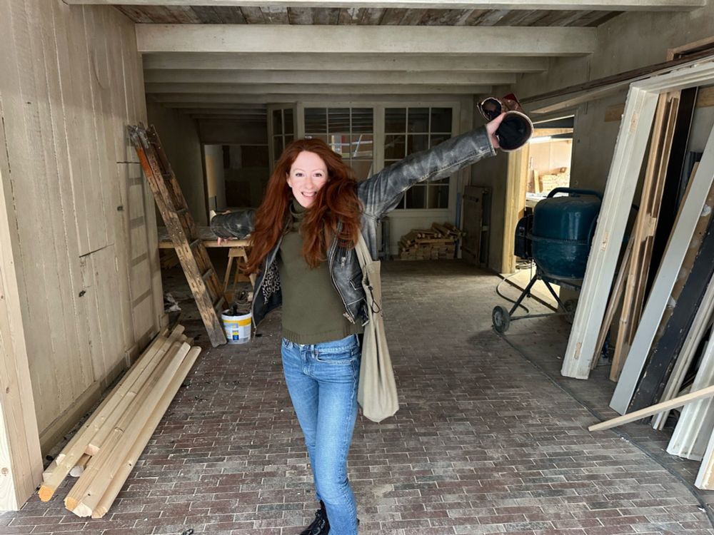 Roxane Duran, who plays Madeline, arms spread, with her baseball cap in her hand, poses in the space that will become Madeline’s Atelier. She is wearing light-wash jeans, a green shirt, and a black jacket. 