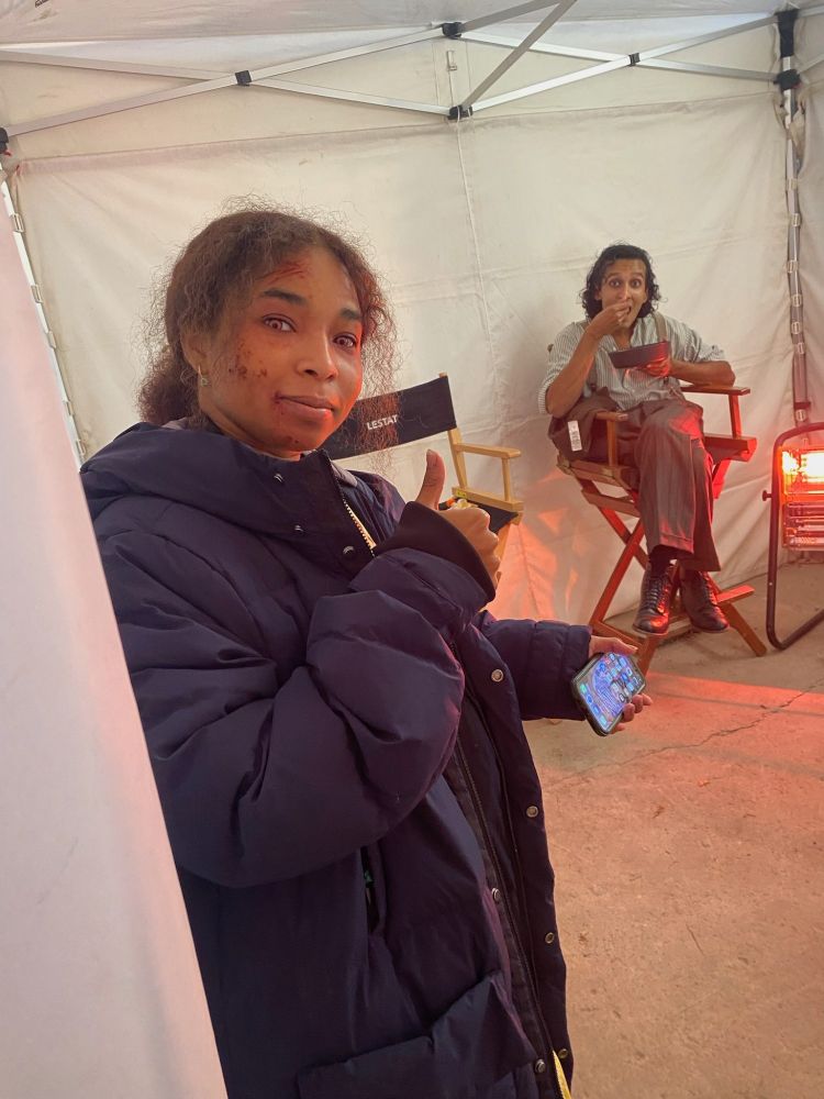 Delainey Hayles stands in a parka at the entrance of a white pop-up tent, holding her phone in one hand and a thumbs up with the other. In the background, Assad Zaman, in costume as Armand, sits in a director’s chair, mid-bite, next to a heater. 
