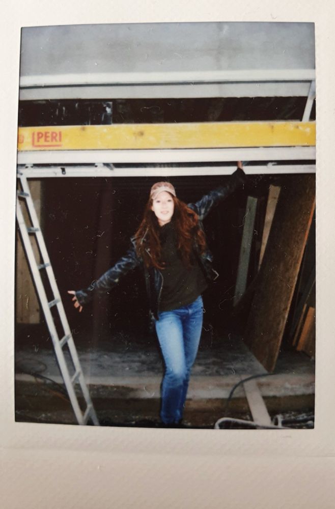 A Polaroid photo of Roxane Duran, who plays Madeline, arms spread with one high and one low, in front of the largely empty build for Madeline’s Atelier. She is wearing light-wash jeans, a hat, a green shirt, and a black jacket. 