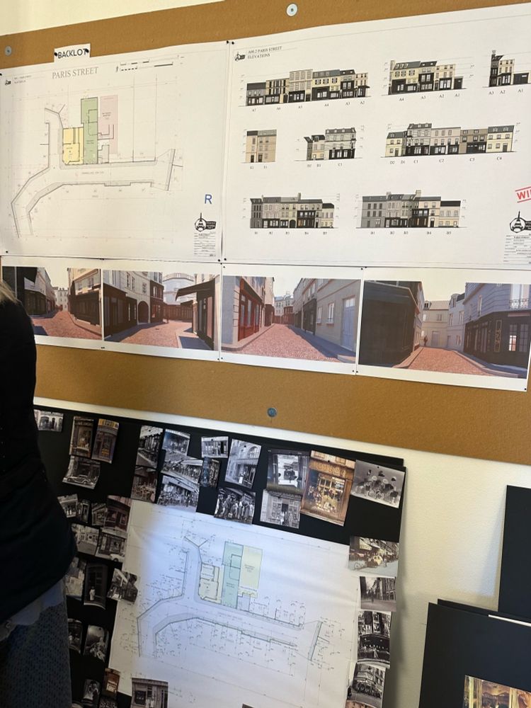 A corkboard with a blueprint layout of the Parisian street backlot, digital mockups of the exterior of the stores, and digital renderings of what it will hopefully look like if one were to walk down the backlot street. 