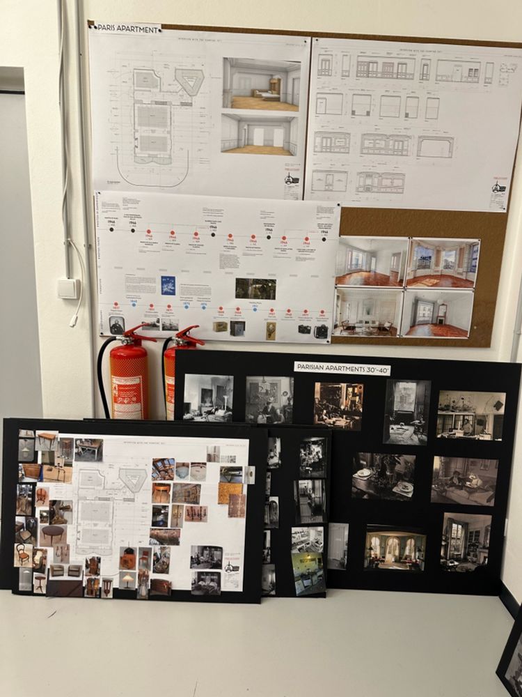 There is a corkboard with three large pieces of white paper and four photos of Parisian apartment interiors. A slip of paper above the top left says, “Paris Apartment.” Under that, there is a blueprint of Louis and Claudia’s apartment build. The paper below is a timeline history of photography from 1857 to 1946. The paper on the top right has more specific build specs. There are two fire extinguishers under the corkboard. Several black design boards are overlapping on the floor against the wall. The board closest to the camera has another blueprint of Louis and Claudia’s apartment, surrounded by photos of furniture and decoration inspiration. The other boards say “Parisian Apartments 30-40s, with photos of apartment interiors from the time.
