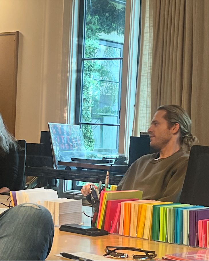 Sam Reid holds a water bottle at the Writer's Room writing table. Exec Producer Hannah Moscovitch sits next to him off-camera. Ryan Kattner's leg is crossed on the other side of the table in front of lines of colorful and white notecards.