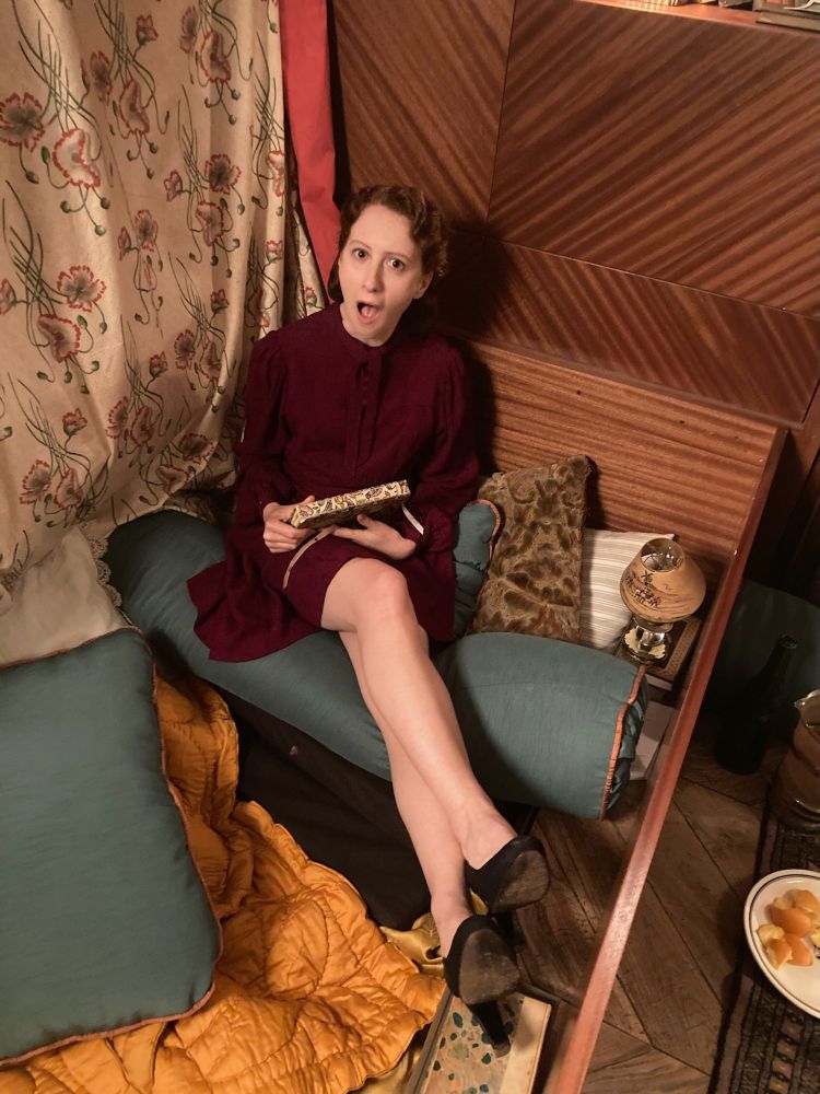Roxane Duran, in costume as Madeline, sitting in Louis’s coffin in Louis and Claudia’s apartment. She is resting on a green oblong pillow with one of Claudia’s diaries in her hands, looking up at the camera, legs crossed, making a shocked face.