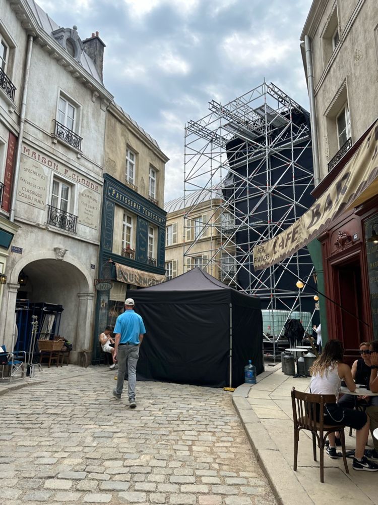 Our Parisian Street backlot, with a black popup tent and a black tented structure, is surrounded by scaffolding at the end of the street. “Stores” on the street include “Patisserie Toussaint," “Maison P. Cloude,” and “Café Bar.” A couple of crew members sit at the “café” while a crew member in a bright blue shirt crosses the street.
