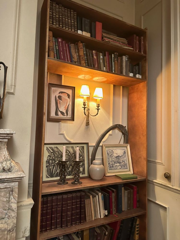 The bookshelf in Louis and Claudia’s apartment is filled with vintage books, largely without visible titles. There are two shelves of just books at the top, then an open section where we can see the white wainscotting of the room, a lamp designed to look like two candles and one framed painting of 2 people embracing is hung next to the lamp. There are two other framed pieces of art, one of which appears to be a charcoal drawing of a group of men and the other a landscape. Beneath the open section are two more rows of books. 