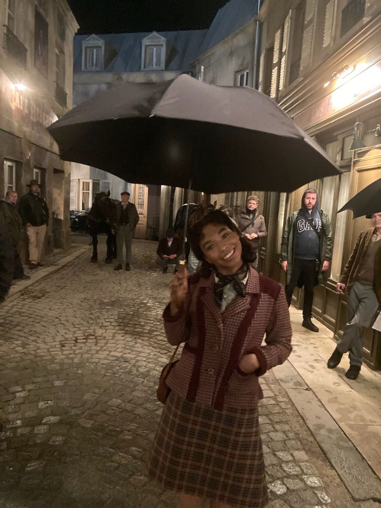 Delainey Hayles, as Claudia, stands on the studio backlot version of a “Paris Street”, with an open umbrella in her right hand. She’s wearing a green and yellow tartan skirt, a jacket with a red gingham pattern and solid red blocks, a brown purse over her shoulder, and a dark green ascot around her neck. Her free hand is in her pocket. Several crew members are around in the backyard, with a few costumed extras and a horse.