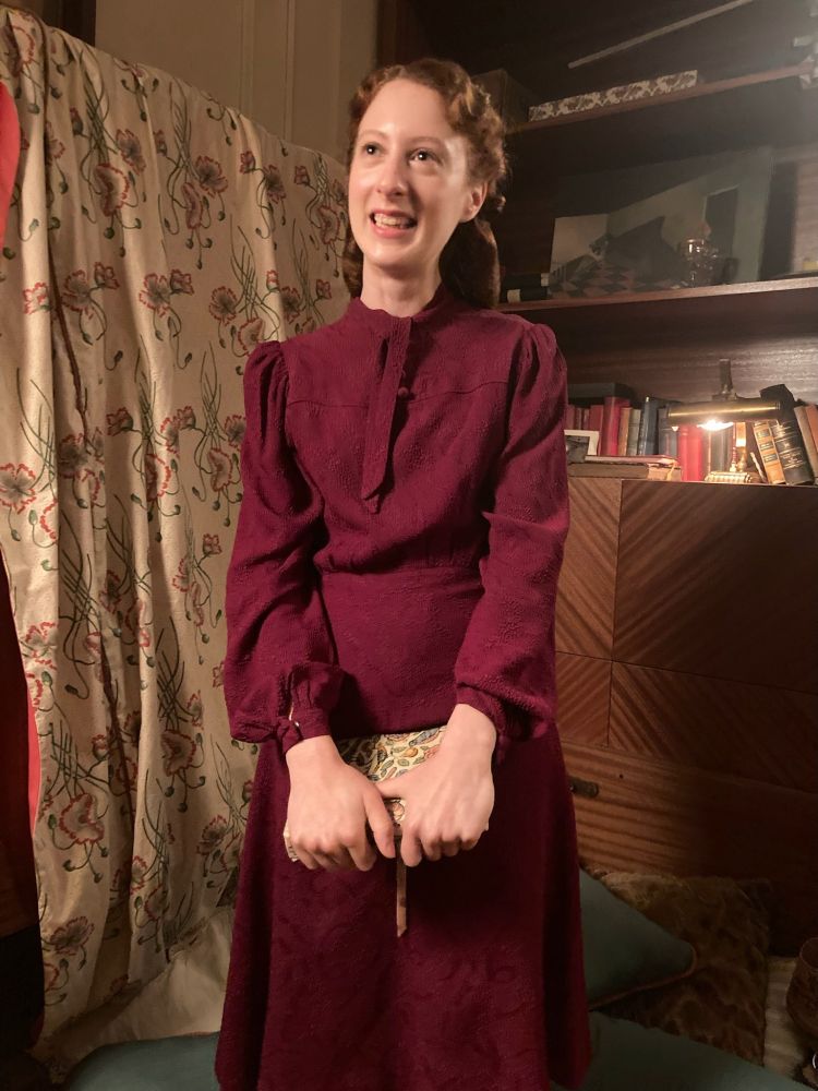 Roxane Duran, in costume as Madeline, stands in Louis and Claudia’s apartment. She holds one of Claudia’s diaries in her hands. She is looking up and to the right of the camera, smiling. 