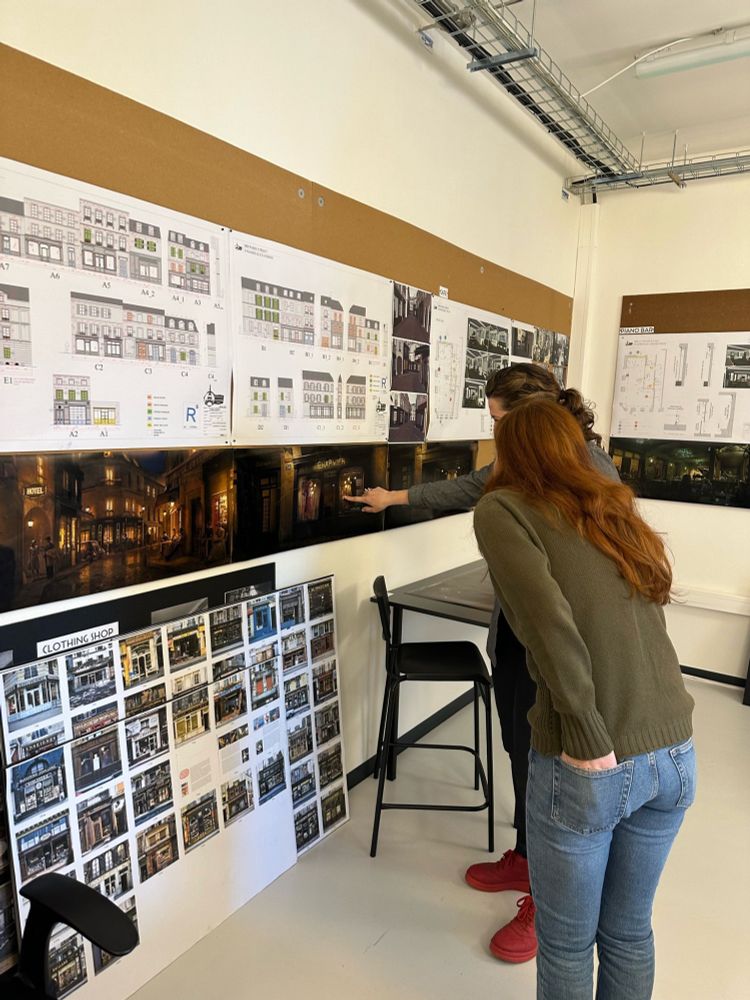 Roxane Duran, who plays Madeline, stands in the art department offices, leaning in to see the design boards for Madeline’s Atelier. Production Designer Mara LePere-Schloop is on her right, obscured mainly by Roxane, but her bright red shoes are visible. Roxane is wearing light-wash jeans, a green shirt, and a black jacket. 