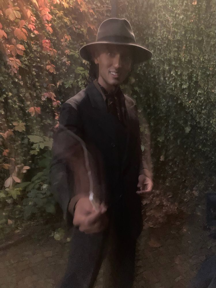 Assad Zaman, as Armand, stands in a park against a wall of ivy, offering the camera a blurry cigarette. He is wearing a purplish brown suit with an ascot and hat.