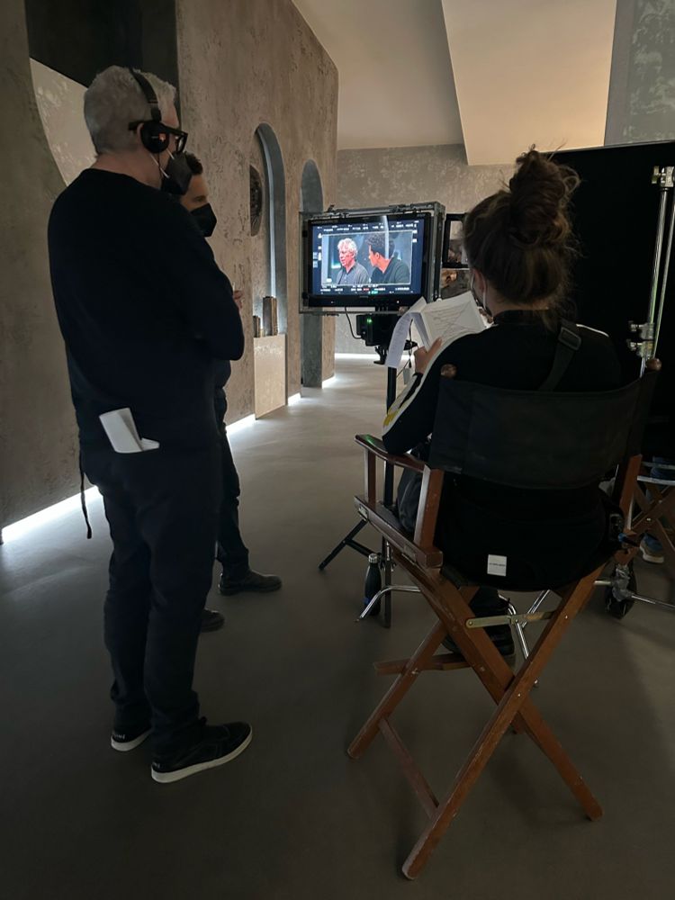 In the middle of the photograph is a monitor, which shows a medium shot of Jacob Anderson and Eric Bogosian in costume as Louis and Molloy, respectively, sitting next to each other. On the right side of the photo, Executive Producer Hannah Moscovitch is sitting on a director’s chair watching the monitor. On the left side, Rolin Jones (Showrunner) and another crew member stand and watch the monitor.