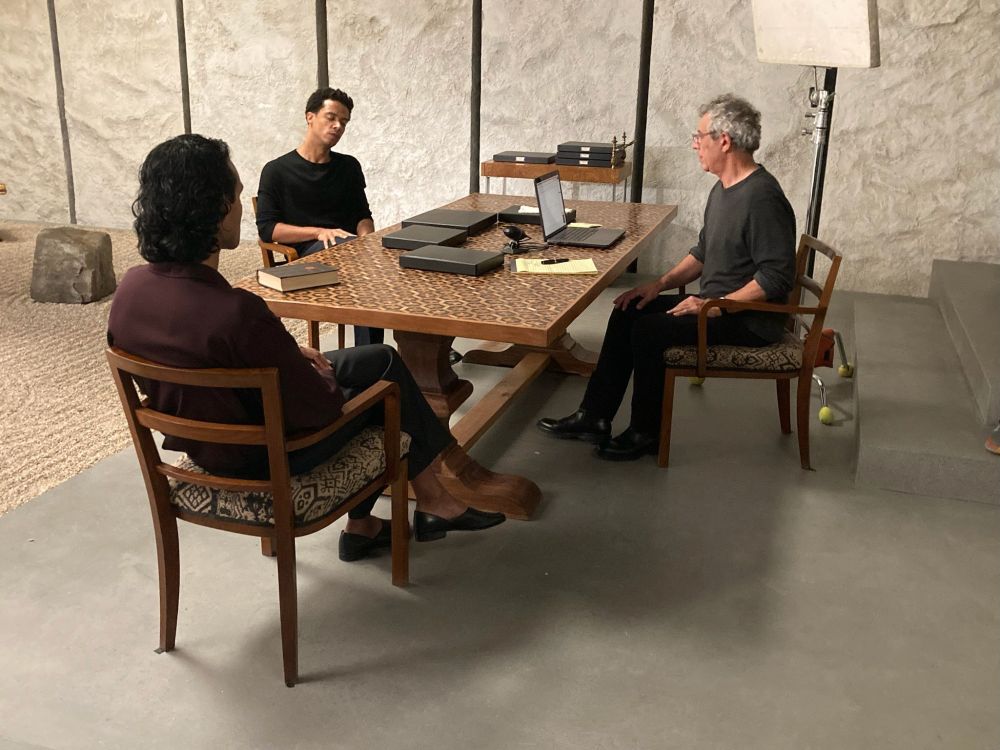 Eric Bogosian as Molloy (on the right), Jacob Anderson as Louis (on the left), and Assad Zaman as Armand (on Jacob’s left) are sitting by a big wooden table in the Dubai Sundial Room. Molloy has his laptop open in front of him and black document-size boxes are laid on the table. A production light is over Eric’s shoulder.