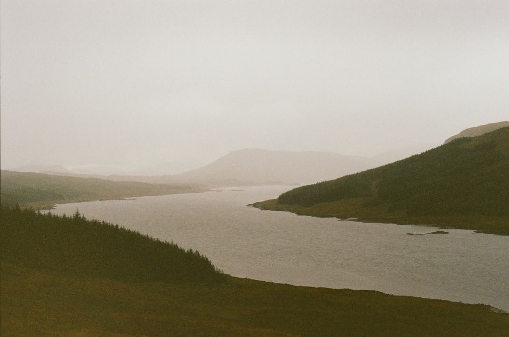 A grainy color photograph of a foggy Scotland landscape with a few small mountains and river.