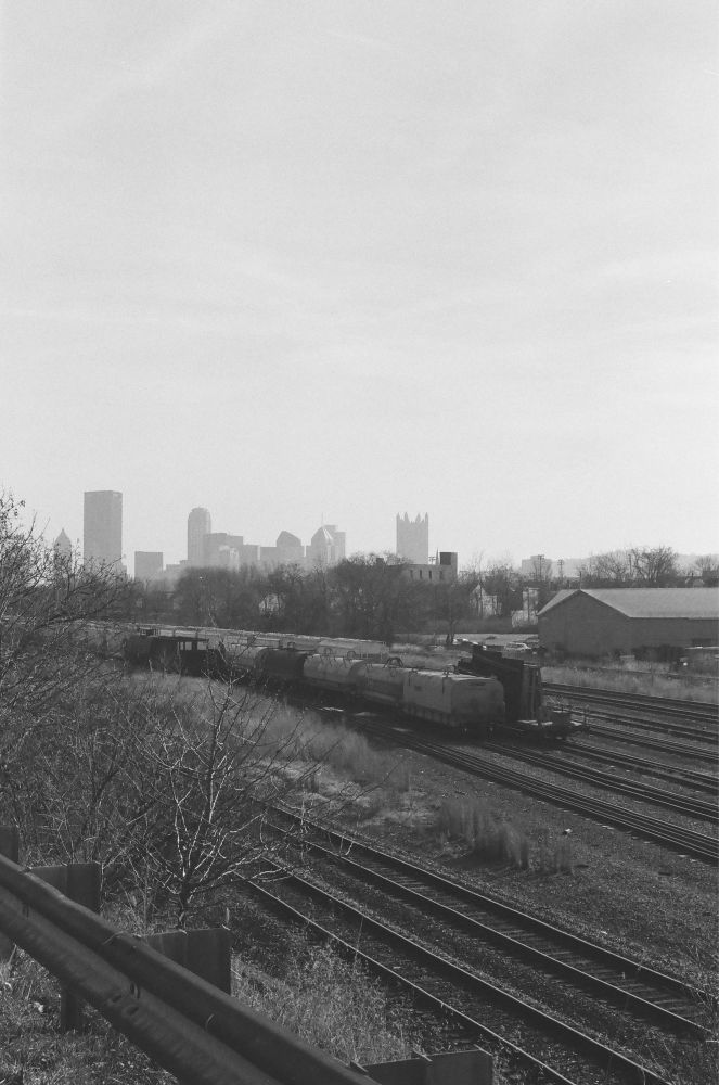 A black and white vertical image of stopped trains in the foreground and the city of Pittsburgh in the background.
