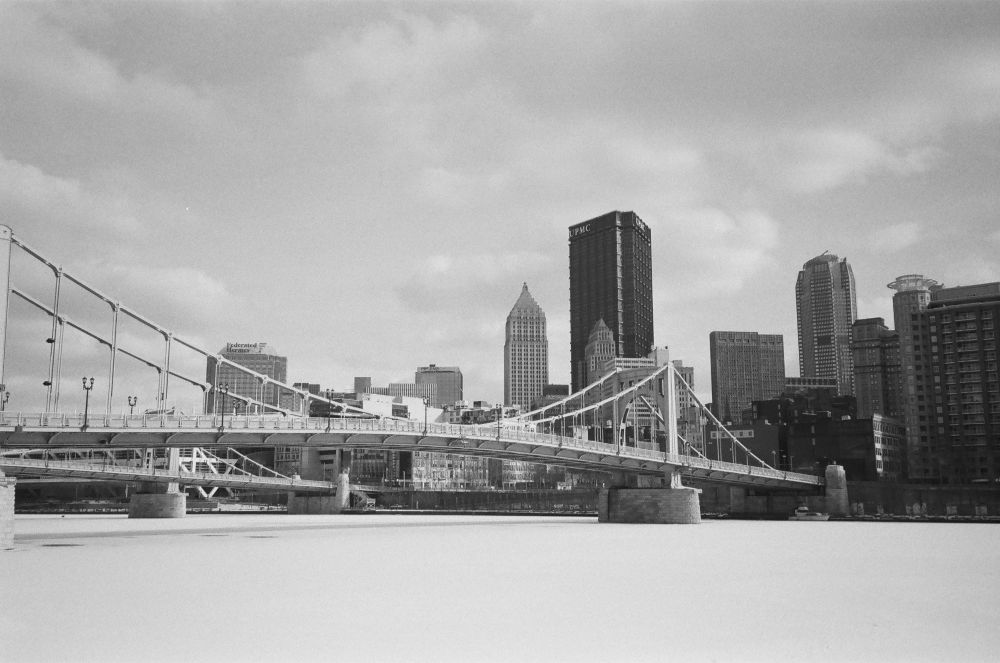 A black and white photo of the Pittsburgh skyline with the frozen, snow-covered river in the foreground.