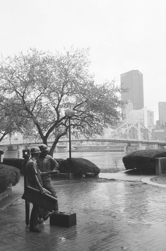 A black and white photo of a statue of surveyors in the foreground and the Pittsburgh skyline in the background during a rainstorm.