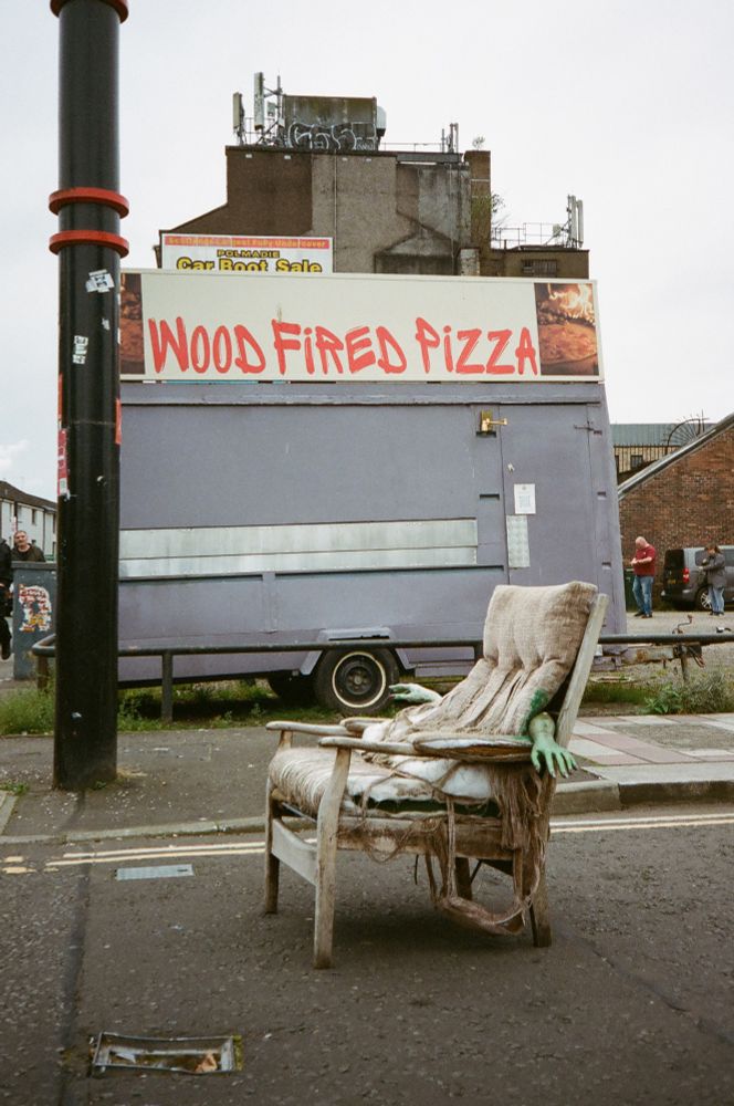 Street art of mannequin hands coming out of an old chair with a wood fired pizza truck in the background.