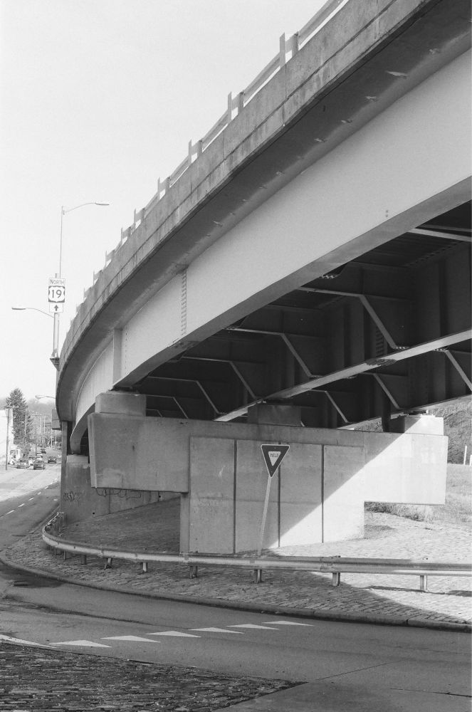 A black and white vertical image of a yield sign under a curving overpass.