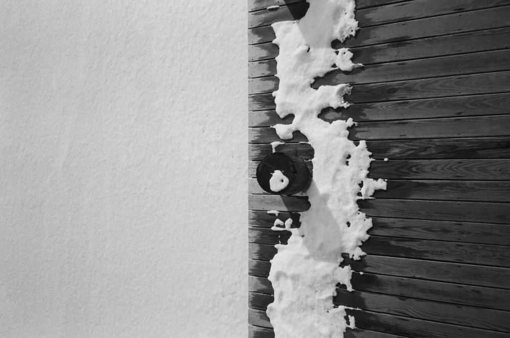 A top down, black and white photo of a wooden dock with snow on it against a frozen, snow-covered river.