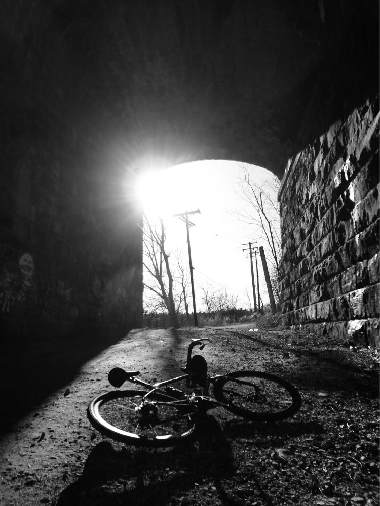 A black and white image of a bicycle laying down on a gravel road in a tunnel with a sun flare peeking through.