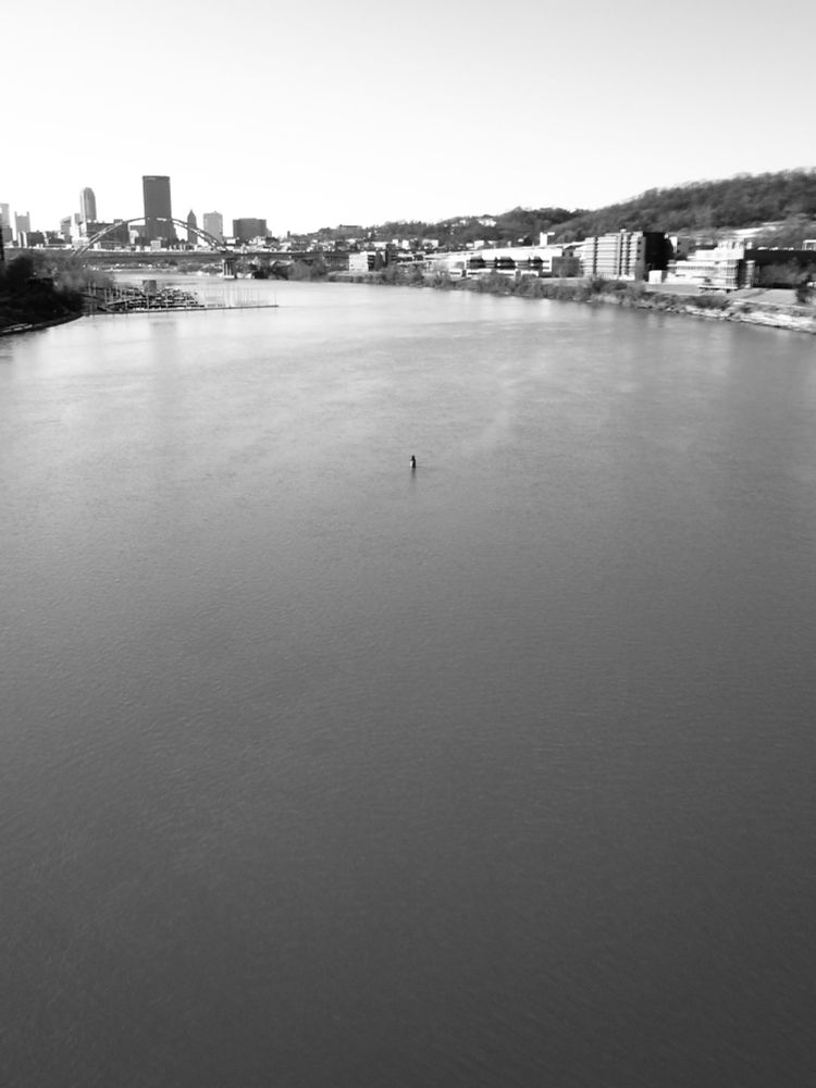 A black and white image of a river in the foreground and the city of Pittsburgh far in the background.