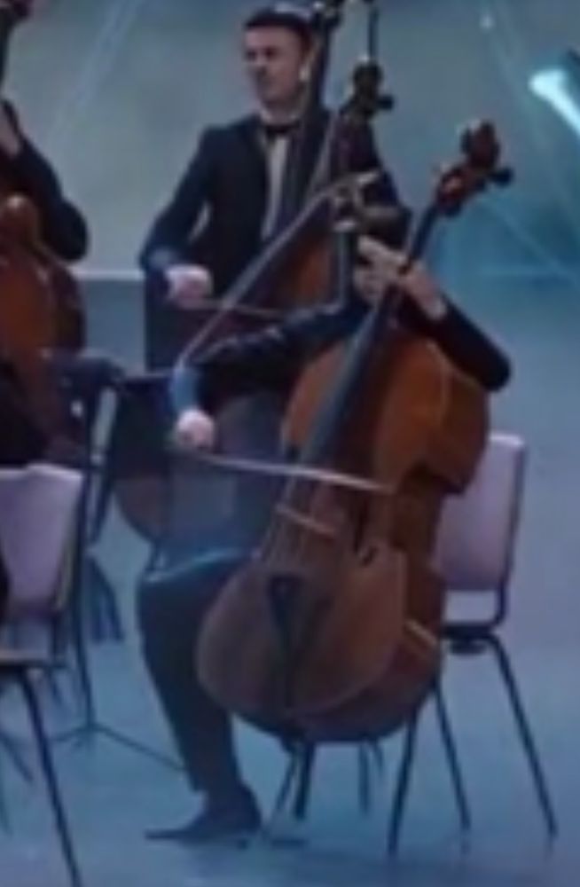 Zoom-in on OPs screengrab of AI tribute video to Captain James T which, as usual, defies description, but I will try. What seems to be a concert in heaven to welcome Cap’n is zoomed in, past the angel choir and to stage left (our right) into the cello/bass section of the orchestra. In poor resolution, one cellist is revealed to have one leg; more importantly, the musician is dressed in what can be a be described as a ninja top, obscuring all but a grimacing mouth. Their head is perfectly sliced to about a 35 degree angle, and this perfect sphere is the base of some sort of crane/pulley that is attached to his bowing hand. Their fretting hand is either so elongated that it is easier for the player to slide pitch to pitch, or, just as likely, their hand has been stabbed through by the instrument’s neck, with the headstock and tuning machines reassembled after bisecting this very large hand.

Also the fretboard seems to taper off with paint drippings and the bow is very uneven and wobbly.

A man is behind this player with an impossibly cinched jacket and some similar device from the headstock to his head.