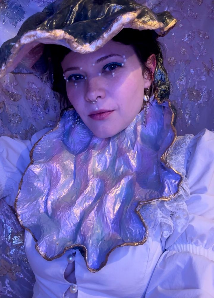 A dark haired afab person adorned with face pearls is wearing a paper mache oyster shell on their head with a white puffy top and a tiny bit of side boob 