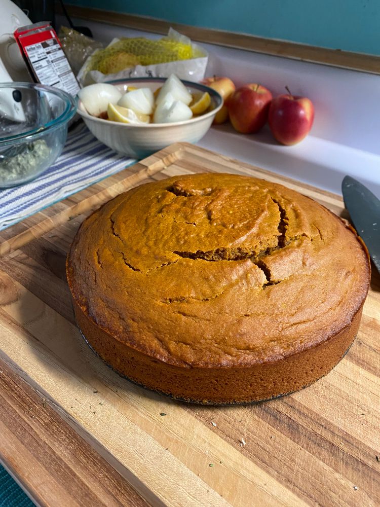 Olive oil pumpkin cake, shown resting on a wooden cutting board. Cracks have opened up on the top of the golden-brown cake, but they will eventually be covered with salted maple cream.