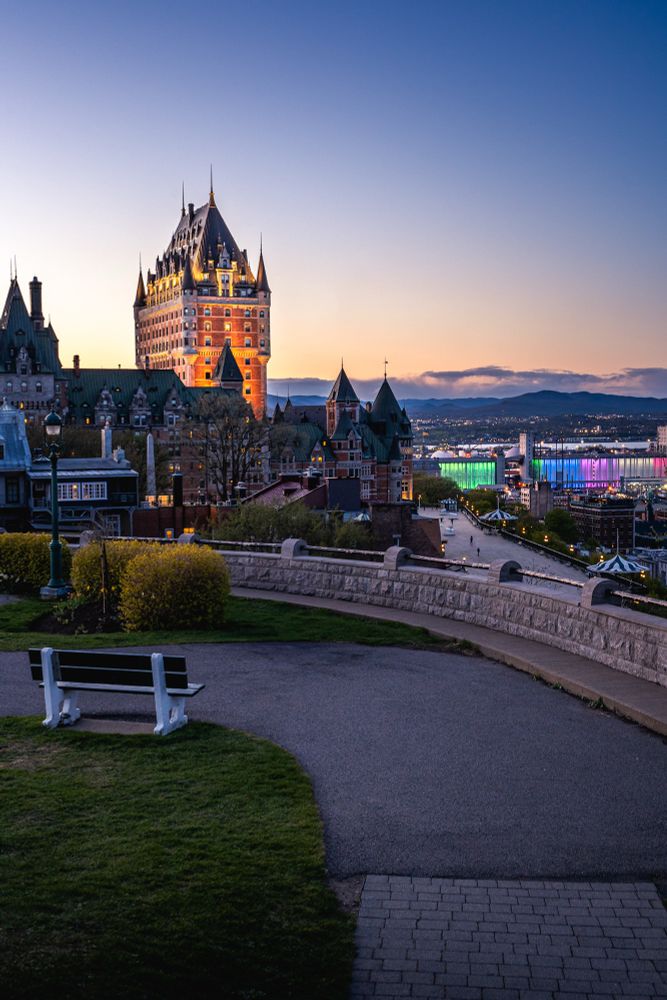 Illuminated castle-like building at dusk with a cityscape and colorful lights in the background.