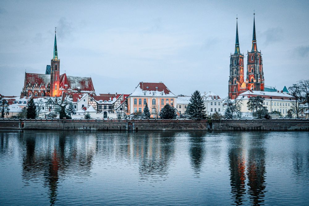 Snow-covered cityscape with historic buildings and church spires reflected in a calm river at dusk.