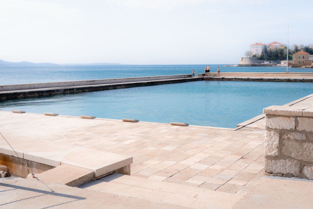 An expansive outdoor swimming pool with shimmering blue water sits adjacent to a tranquil sea or ocean. The pool is bordered by a spacious, tiled deck, with a few people casually gathered on the opposite side, adding a hint of activity to the serene setting. In the distance, there’s a charming coastal town featuring whitewashed buildings and a notable structure perched atop a small hill. The backdrop also reveals gentle waves of the sea with distant islands or landmasses, enhancing the picturesque blend of nature and human architecture.