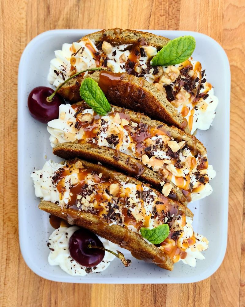 3 tacos lined up on a dark plate. Banana bread shell, whipped white chocolate ganache, salted bourbon caramel, toasted walnuts, dark chocolate. Cherries with whipped cream are added to the plate.