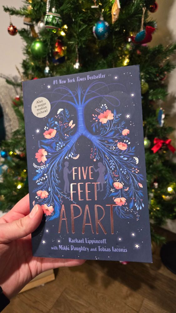 Paperback copy of Five Feet Apart by Rachael Lippincott, Mikki Daughtry and Tobias Iaconis held up in front of the Christmas tree.