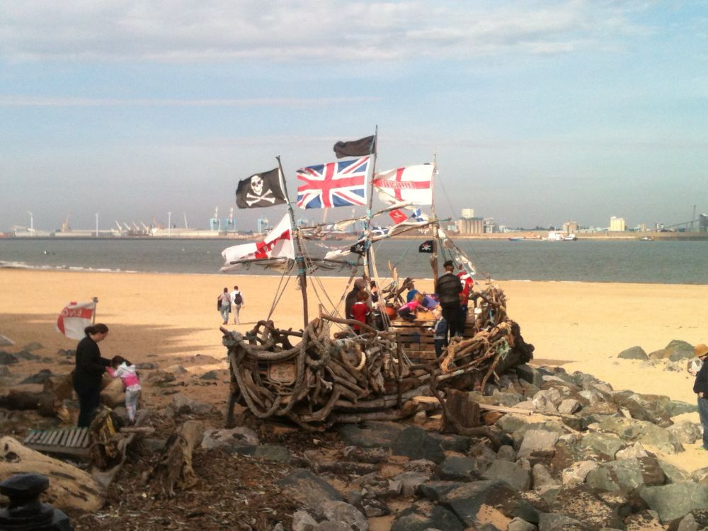 A makeshift pirate ship on the shore of the Mersey River in Wallasey, UK, looking toward Liverpool. There are a few people on and around the ship made of things washed ashore, and adorned with flags. 