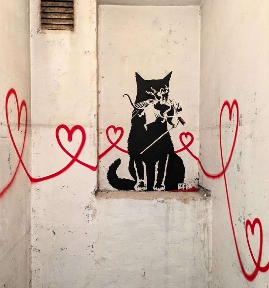 Streetartwall.  A stencil mural with a cat, a rat and red hearts was sprayed into a white wall niche. The black cat by artist "Tabby" sits in the niche and has a rat in the style of "Banky" in its mouth. The Banky rat is holding a stick with a paint roller. From the cat, curved and interconnected hearts extend beyond the two walls into another room of an exhibition.
Info: The TABBY Vienna exhibition at Hotel Kunst BTQ will take place from November 29 to December 1. 2024.
Tabby has freely designed the walls of the exhibition rooms. Admission is free but only open via guest list in the Website.