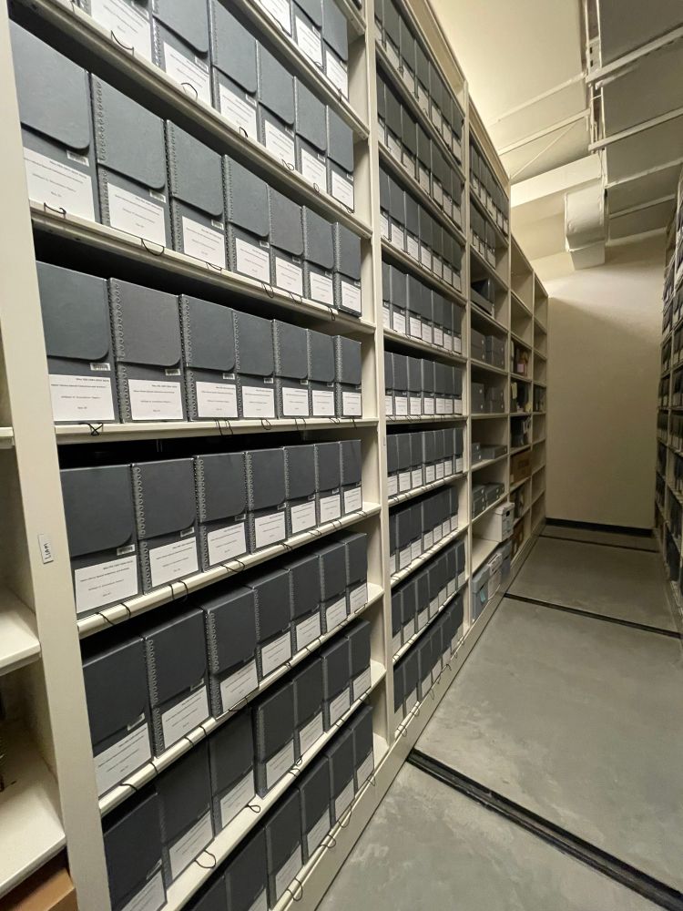 Several shelves of neatly arranged gray, archival boxes with white labels. 